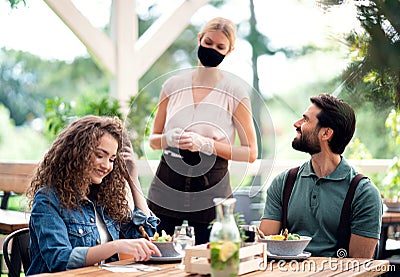 Waitress with face mask serving happy couple outdoors on terrace restaurant. Stock Photo