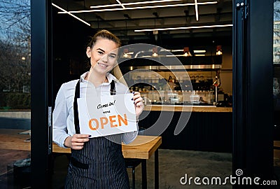 Waitress wearing apron standing at restaurant entrance. Woman attend new customers in her coffee shop. Happy woman showing open Stock Photo