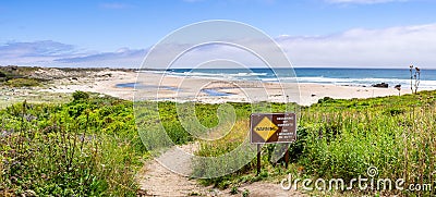 Walking path going through green shrubs towards a sandy beach; sign warning of Recurring Rip Currents posted on the trail; Gazos Stock Photo
