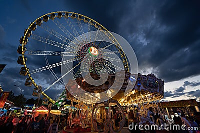 Walking Tourists close to the carousel with the Ferris Wheel in the Background on a cloudy sunset Editorial Stock Photo