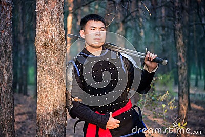 Warrior in black armor with a sword in his hands is leaning against a tree in a gloomy enchanted, damned forest Stock Photo