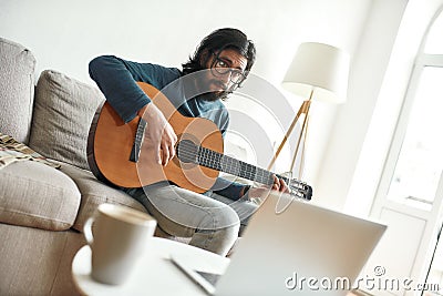 Watching video tutorial. Young man sitting on sofa at home and learning guitar online. Sitting on sofa at home and Stock Photo