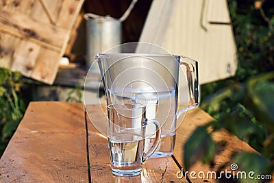 Water filter jug and a transparent cup of water with a village well on the background Stock Photo