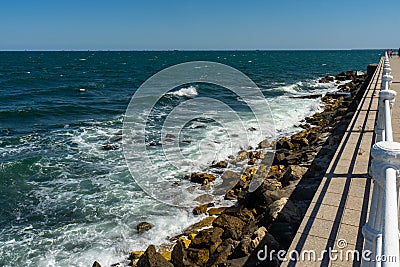 The waves of the Black Sea crash against the rock base of the pedestrian promenade in Constanta, bordered by a white railing. Stock Photo