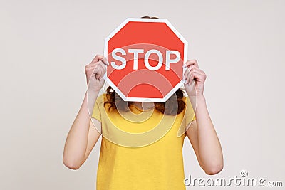 Way prohibited! Portrait of anonymous girl in yellow casual T-shirt covering face with stop symbol, holding red traffic sign, Stock Photo