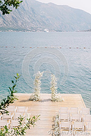 Wedding arch decorated with white flowers stands on a pier in front of rows of white chairs Stock Photo