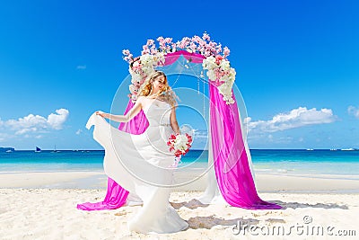 Wedding ceremony on a tropical beach in purple. Happy blond bride with wedding bouquet under the arch decorated with flowers on t Stock Photo