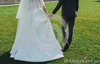 Wedding couple is holding hands and walking outside in the bright green lawn. Bride in white elegant wedding dress with train is Stock Photo