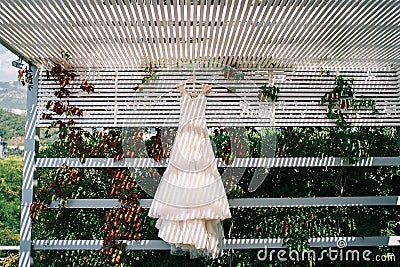 Wedding dress hangs on a hanger in the striped shade of a wooden slatted canopy in the garden Stock Photo