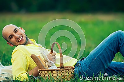 Weekend picnic concept. Portrait of a young handsome bald man in trendy clothes lying on green grass in the park. Picnic basket Stock Photo