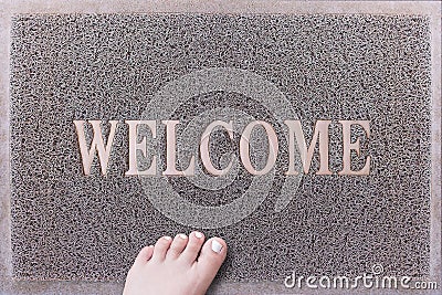 Welcome Door Mat With Female Foot. Friendly Grey Door Mat Closeup with Bare Woman Foot Standing. Welcome Carpet. Stock Photo