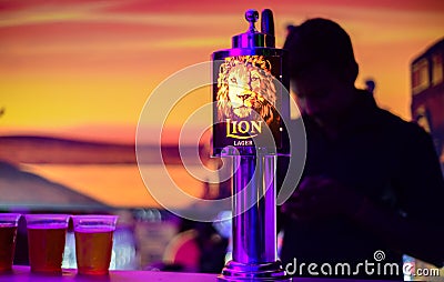 Lion lager backlit logo on a liquor pub table and 3 beer cups. Silhouetted bartender in the Editorial Stock Photo