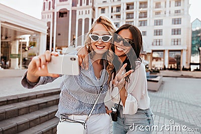 Well-dressed blonde lady making selfie with sister. Outdoor shot of romantic girls having fun in sunny spring day.. Stock Photo
