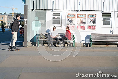 People seated and walking by old wharf shed with roller-blade hire advertising posted on wall on city waterfront area Editorial Stock Photo