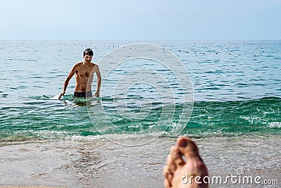 A wet, handsome dark-haired man emerges from the water on a Mediterranean beach in Alanya, Turkey. Blurred female foot against the Stock Photo