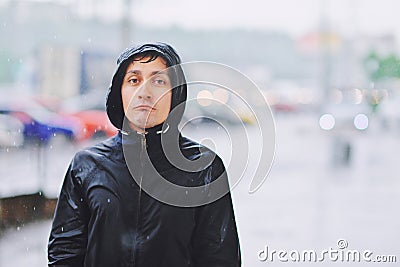 Wet young man in raincoat with unhappy face walks along the street during heavy rain, close-up. Autumn bad stormy weather concept Stock Photo