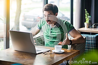 What do you want? Young angry businessman in green t-shirt sitting, working, looking and screaming at laptop screen on video call Stock Photo