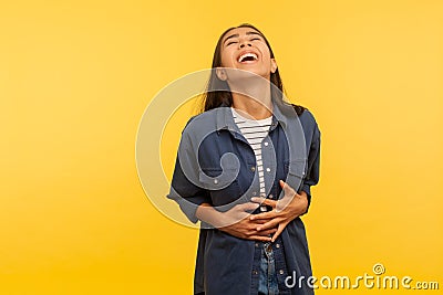 What a funny joke! Portrait of amused carefree girl in denim shirt cracking up with hilarious laughter, holding her stomach Stock Photo