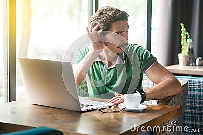 What? I can`t hear you. Young businessman in green t-shirt sitting and try to listen something on laptop or video call Stock Photo