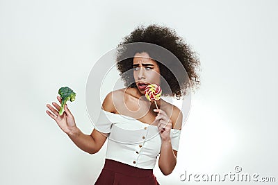 What to choose Frustrated afro american woman is facing a choice of healthy broccoli or sweet lollipop and looking at Stock Photo