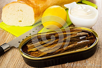 Baguette, lemon on cutting board, knife, bowl with mayonnaise, opened jar with sprats on wooden table Stock Photo