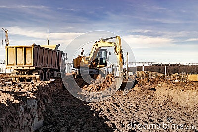 A wheeled excavator loads a dump truck with soil and sand. An excavator with a high-raised bucket against a cloudy sky View from Editorial Stock Photo