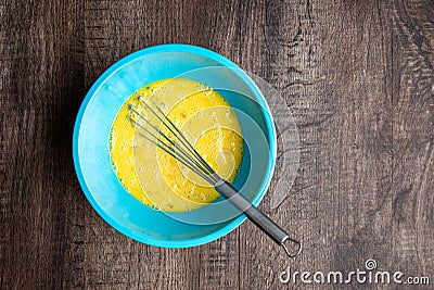 Whipped eggs in blue mixing bowl, metal whisk, wood table Stock Photo