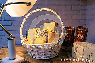 A white basket filled with various cheeses against a brick wall. Aged cheese is illuminated by a table lamp Stock Photo