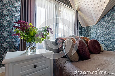 White bed in bedroom, bedside table with standing bouquet of lilacs Stock Photo