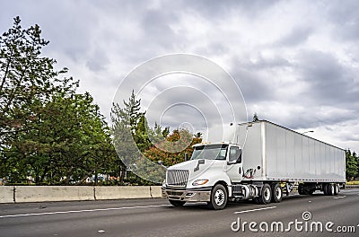White big rig day cab semi truck with roof spoiler transporting goods in dry van semi trailer driving on the multiline highway Stock Photo