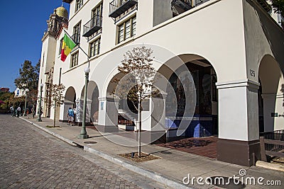 A white building with brown trim and a colorful wall mural and a red, green and yellow flag with people walking Editorial Stock Photo