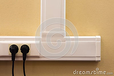 White cable channel with electrical outlets on a yellow wall, where two black plugs are inserted. Stock Photo