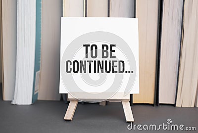 White card with text To be continued stands on the desk against the background of books stacked Stock Photo