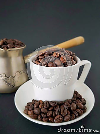 White coffee cup and saucer; coffee beans scattered on the table. In the background is a cezve. Black background. Stock Photo