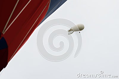 White coloured blimp, hot air airship in grey sky in rainy day nearby with vivid red Hot Air Balloon. Inflatable Stock Photo