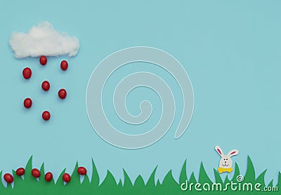 White cotton cloud with the rain of small red Easter eggs falling on green grass and Easter bunny on blue background. Happy Easter Stock Photo