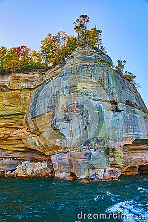 White cresting waves in teal lake waters beneath pictured rocks cliff face with fall trees Stock Photo