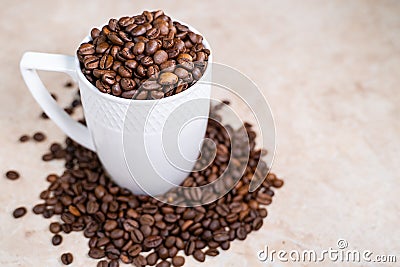 A white cup filled with roasted cinnamon coffee beans standing on brown marble surrounded by coffee beans Stock Photo