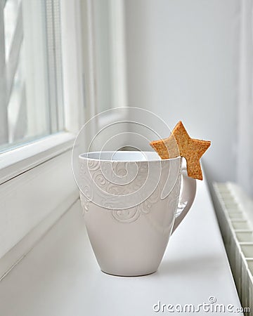 A white cup of tea or a cup of coffee with a cookie in the shape of a star by the window. Stock Photo