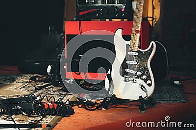 A white electric guitar and a red amplifier onstage with electrical audio cables and sound effects pedals Stock Photo