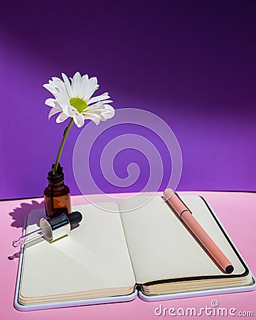 A white flower, a bottle with an eyedropper and a notebook on the background of a purple wall. Stock Photo