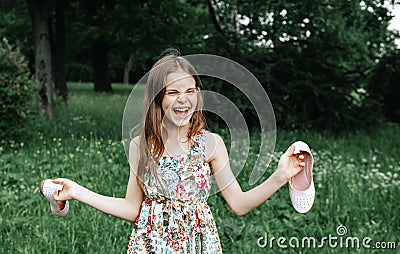 Girl 10 years old in a dress holding shoes in her hands and laughing in a clearing in nature rejoicing in nature Stock Photo