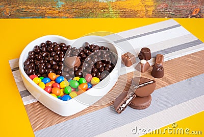 White heart-shaped bowl filled with brown and colorful chocolates, hazelnuts and almonds covered in chocolate Stock Photo