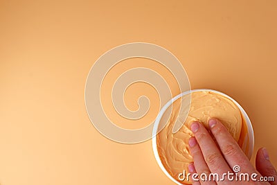 In a White jar, peach-colored body cream, woman's hand takes cream. Orange background. Copy space Stock Photo