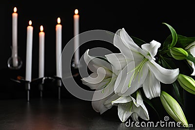 White lilies and blurred burning candles on table in darkness, closeup with space for text. Stock Photo