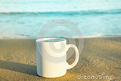 White Mock Up Mug with Empty Space for Artwork Text Standing on Beach Sand. Turquoise Blue Sea Stock Photo