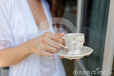 White mug with a pattern with hot coffee inside the in the hands of women in Bathrobe standing on the balcony of her bedroom. Woma Stock Photo
