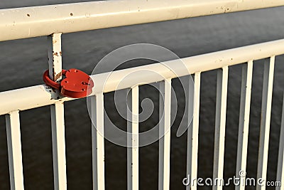 The white painted railing of the embankment of the river. With a red lock in the shape of a heart, mounted on a metal pipe. Stock Photo