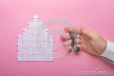 White puzzle. House shape puzzle. The concept of rent, mortgage. Hand holding keys. Pink background. Top view Stock Photo