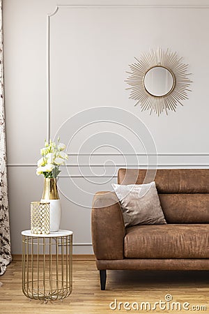 White roses in a vase on the stylish table next to brown leather sofa in light living room Stock Photo